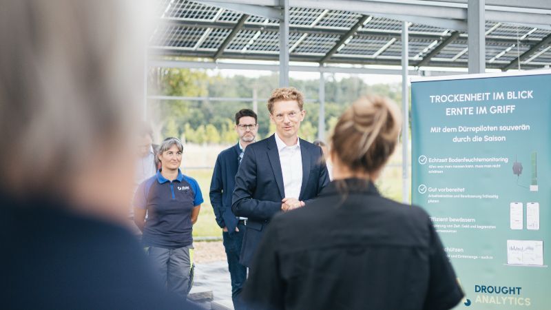 Dr. Georg Schaumann von SenseUp steht im Mittelpunkt einer kleinen Gruppe. Er trägt einen dunklen Anzug und lächelt leicht in Richtung Kamera. Neben ihm sind weitere Personen zu sehen, darunter eine Frau im blauen Poloshirt. Rechts im Bild steht ein Roll-up von Drought Analytics mit Hinweisen zum Echtzeit-Bodenfeuchtemonitoring und effizienter Bewässerung. Im Vordergrund ist Ministerin Mona Neubaur von hinten unscharf zu erkennen.