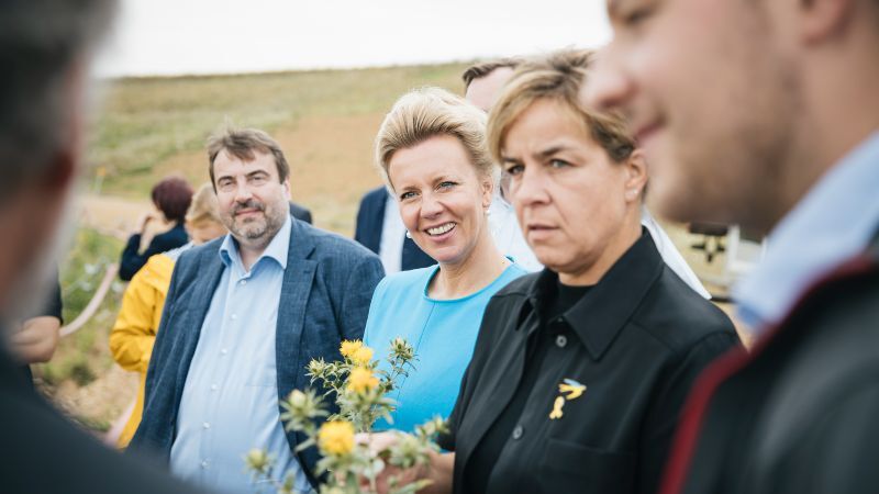 Dr. Corvin Volkholz von Ilco Chemikalien, Ministerin Ina Brandes in einem hellblauen Kleid und Ministerin Mona Neubaur in schwarzer Kleidung stehen gemeinsam mit weiteren Personen im Freien. Ina Brandes lächelt in die Kamera, während Mona Neubaur eine gelbblühende Färberdistel in der Hand hält. Im Hintergrund sind weitere Menschen und ein leicht ansteigendes Feld zu sehen. Die Stimmung wirkt aufmerksam und interessiert.
