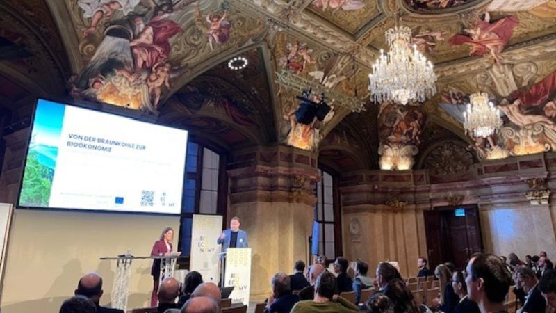 Das Bild zeigt einen herrschaftlichen Saal mit hoher Decke und österreichischen Motiven. Etwas weiter hinten ist eine Bühne zu sehen, auf der Dr. Christian Klar zusammen mit einer Frau vor einer Präsentationsfolie steht.