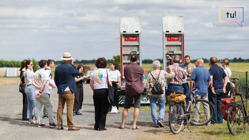 Eine Gruppe von etwa 20 Erwachsenen steht im Freien auf einem Parkplatz oder Feldweg. Sie versammeln sich um zwei geöffnete Ladeflächen eines Lastwagens und hören einer Person zu, die etwas erklärt. Einige Menschen halten Notizblöcke, andere stehen mit Fahrrädern daneben. Im Hintergrund sind Felder und ein bewölkter Himmel zu sehen.