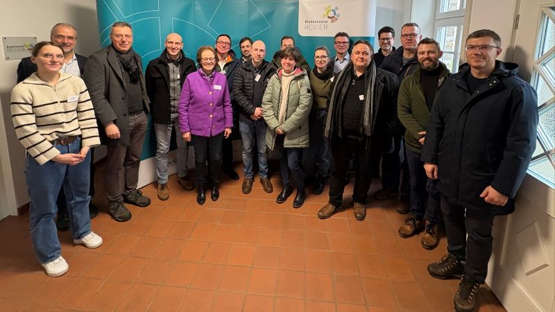 Gruppenfoto im Innenraum des Neesenhofs in Bürgewald: Rund 15 Teilnehmende stehen in zwei Reihen vor einer türkisfarbenen BioökonomieREVIER-Roll-Up-Wand. Sie tragen winterliche Kleidung und Namensschilder und blicken in die Kamera. Anlass war der Austausch zwischen BioökonomieREVIER und Vertreterinnen und Vertretern der LEADER-Region Delitzscher Land am 2. Februar.