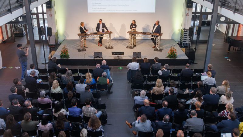 Podiumsdiskussion beim Regionalforum Bioökonomie: Vier Personen stehen auf einer Bühne hinter Stehtischen und diskutieren vor Publikum. Im Saal sitzen zahlreiche Zuhörerinnen und Zuhörer in Reihen, einige verfolgen die Diskussion aufmerksam, eine Person fotografiert. Die Veranstaltung findet in einer modernen Halle mit großer Projektionsfläche im Hintergrund statt.