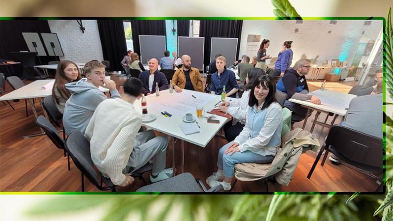 In einem hellen Raum sitzen mehrere Menschen an Tischen in kleinen Gruppen zusammen. Im Vordergrund arbeitet eine Gruppe an einem großen Blatt Papier, auf dem Notizen gemacht werden. Einige Personen schreiben, andere hören zu oder diskutieren. Im Hintergrund sind weitere Gruppen sowie Stellwände und Moderationsmaterial zu sehen. Die Atmosphäre wirkt konzentriert und kooperativ, wie bei einem Workshop oder einer gemeinsamen Arbeitsphase.