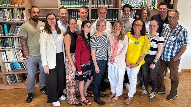 Gruppenfoto während des zweiten Bildungsdialogs von BioökonomieREVIER. 15 Teilnehmende aus Kommune, Bildung und Wirtschaft stehen in zwei Reihen vor Bücherregalen in einem Seminarraum und blicken lächelnd in die Kamera. Sie tragen überwiegend sommerliche Freizeit- oder Business-Casual-Kleidung, einige Namensschilder. Der Raum mit Holzfußboden vermittelt eine helle, freundliche Arbeitsatmosphäre.