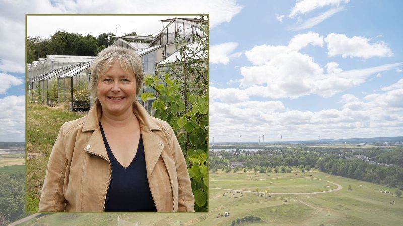 Anke Krüger steht lächelnd vor mehreren Gewächshäusern des Forschungszentrums Jülich. Sie trägt eine beigefarbene Jacke über einem dunklen Oberteil. Im Hintergrund ist ein weiteres Bild einer weiten Landschaft mit Feldern, Grünflächen und Windrädern unter einem blau-weiß bewölkten Himmel zu sehen.