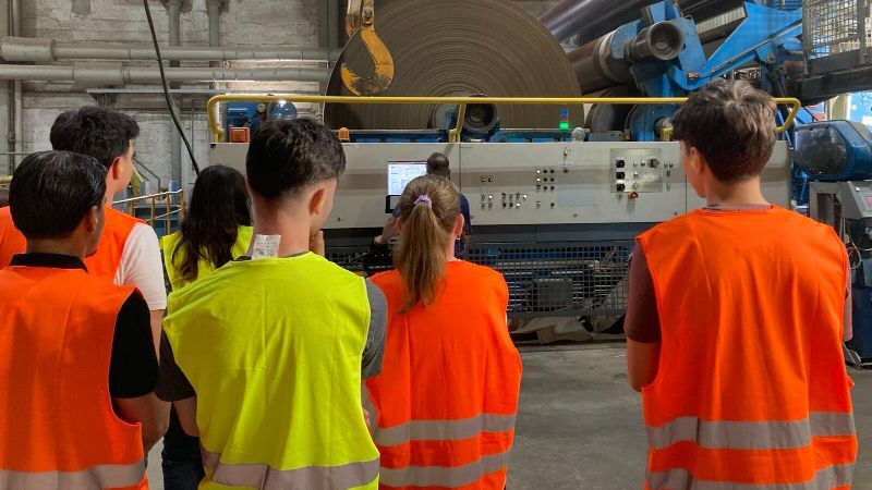 Eine Gruppe Schüler steht in einer Industriehalle vor einer großen Produktionsmaschine. Die Teilnehmenden tragen orangefarbene und gelbe Warnwesten und betrachten die Anlage von hinten. Im Hintergrund sind große Rollen mit aufgewickeltem Material sowie Rohre und weitere technische Komponenten der Produktionsanlage zu sehen.