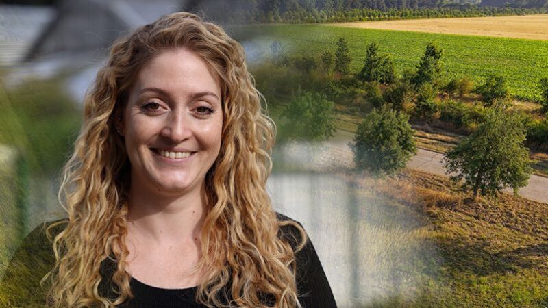 Porträt von Dr. Sabine Dietrich vor einer landwirtschaftlich geprägten Landschaft. Die lächelnde Person mit langen, lockigen, blonden Haaren trägt ein schwarzes Oberteil. Im Hintergrund sind grüne und abgeerntete Felder, Bäume und ein Feldweg zu sehen. Das Porträt geht mit einem weichen Übergang in die Landschaftsaufnahme über.