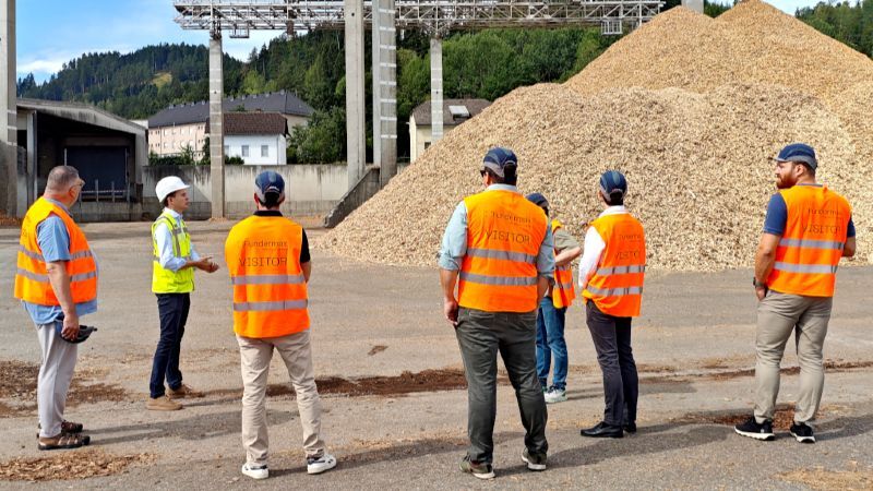 Eine Gruppe von sieben Personen in Warnwesten steht auf einem Betriebsgelände vor einem großen Haufen Holzhackschnitzel. Eine Person mit weißem Schutzhelm spricht zur Gruppe. Im Hintergrund sind Industrieanlagen, Gebäude und bewaldete Hügel zu sehen.