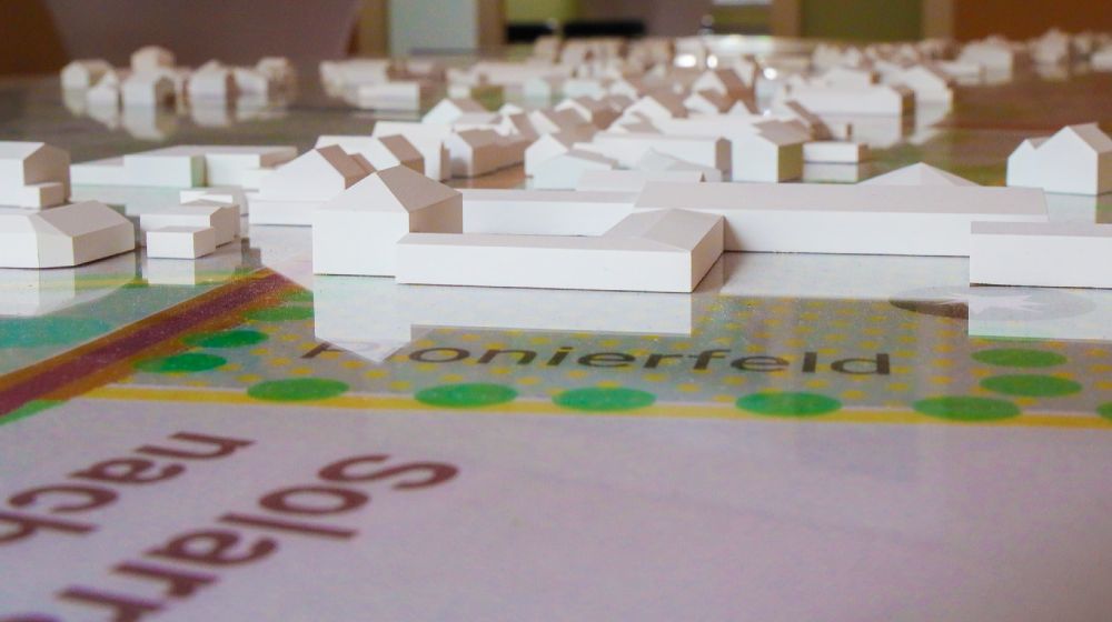 Das Bild zeigt ein maßstabsgetreues Architekturmodell von Merzenich-Bürgewald. Auf einer bedruckten Grundkarte mit Straßen und Grünflächen erheben sich weiße, stilisierte Baukörper, die Wohnhäuser und Gebäude symbolisieren. Die klaren geometrischen Formen deuten eine moderne Siedlungsgestaltung an. Durch die abstrahierte Darstellung werden Strukturen, Wege und Platzierungen sichtbar, die die zukünftige Entwicklung und Planung von Bürgewald  veranschaulichen.