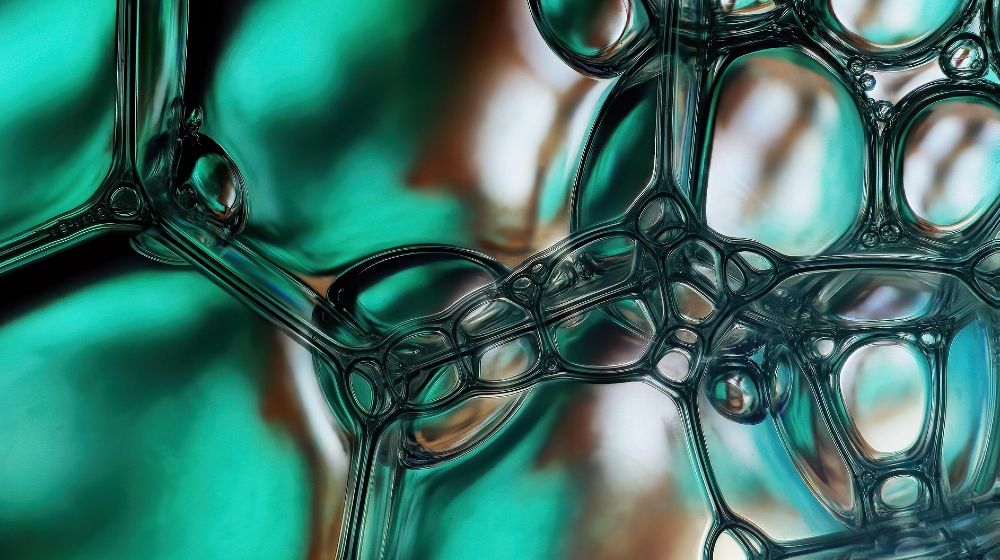 Abstrakte Nahaufnahme schimmernder, transparenter Strukturen, die an Seifenblasen oder flüssiges Glas erinnern. Organisch geformte, miteinander verbundene Blasen bilden ein netzartiges Muster. Die Farben reichen von Türkis und Grün bis zu warmen Braun- und Beigetönen im Hintergrund. Lichtreflexe und weiche Unschärfen erzeugen Tiefe und eine fast mikroskopische, fließende Anmutung.